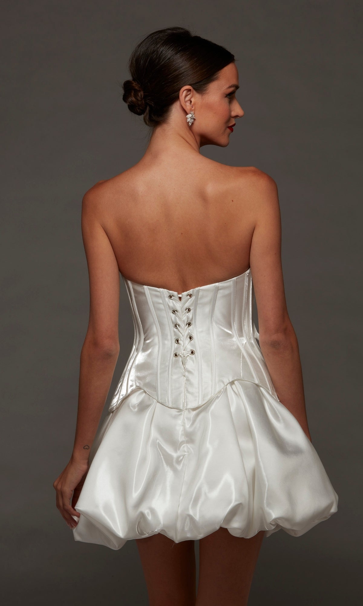A woman showcases the Short Party Dress: Alyce 70108, a strapless corset dress crafted from liquid satin with a dramatic bubble-hem skirt, viewed from the back against a plain background.
